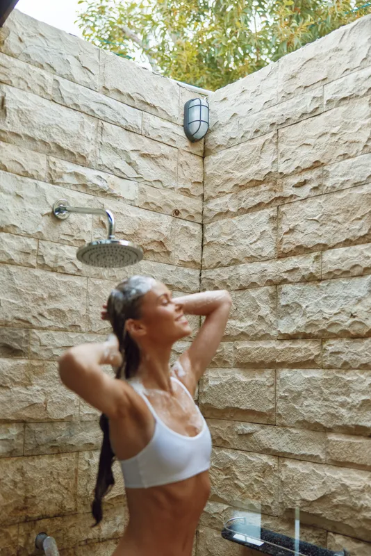 young woman enjoying outdoor shower under natural light with stone wall background and wet hair, feeling relaxed and fresh in summer setting