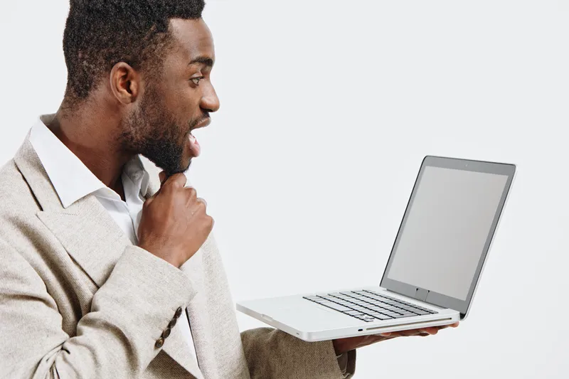 Professional man holding open laptop computer with blank screen, dressed in beige sweater and white shirt, smiling, isolated on gray background. Business, technology, work, education, communication co