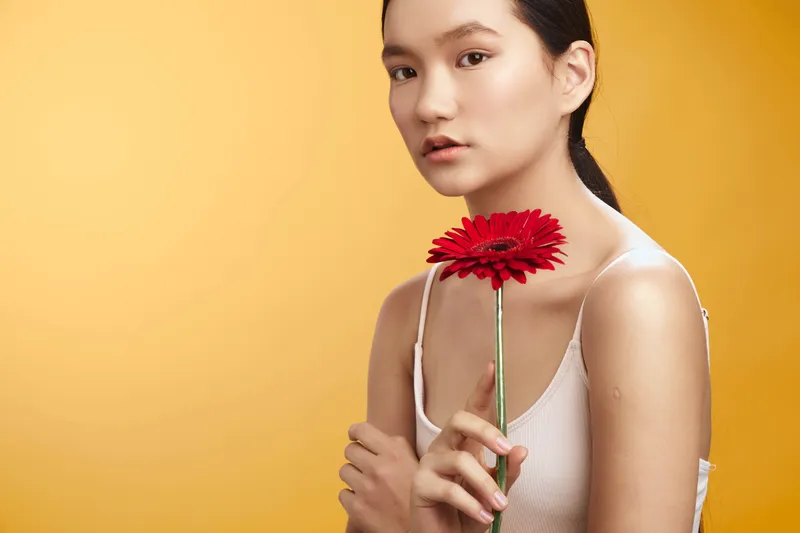 Asian woman holds a red flower beside a warm yellow backdrop, gazing softly at the camera. This studio portrait conveys grace, calm, and natural beauty through gentle lighting and clean composition.