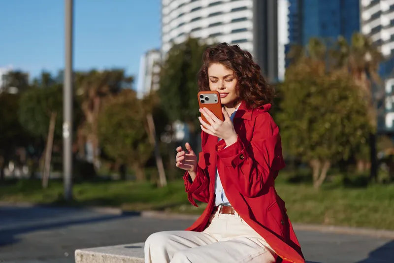 Young woman with curly hair wearing a red coat sits outdoors on a concrete bench while looking at her smartphone on a sunny day.