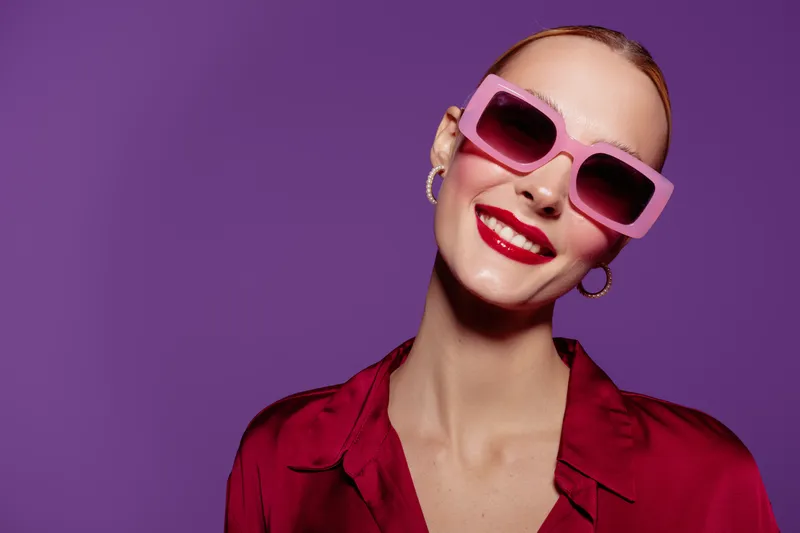 Smiling woman with pink sunglasses and red lipstick poses against purple background. She wears red blouse and gold hoop earrings. Concept of fashion, beauty, and confident style.