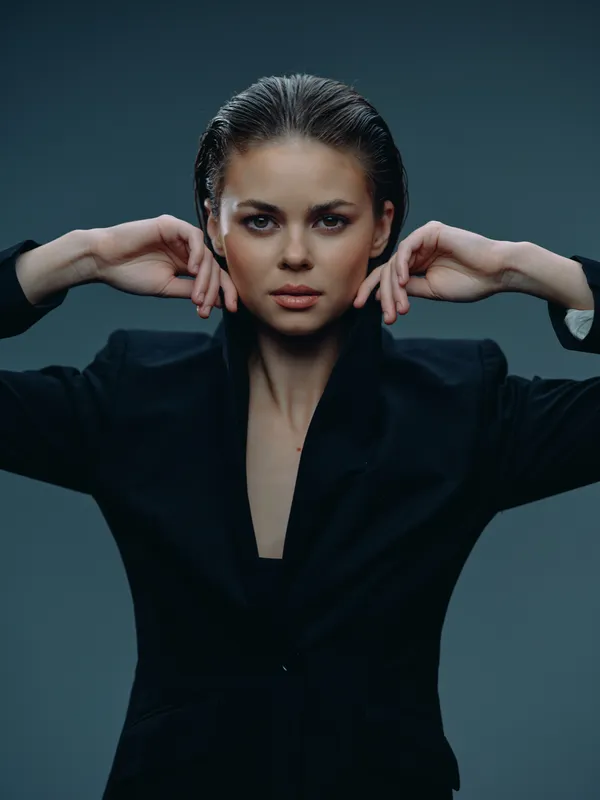 Confident woman in black blazer posing with hands near face, wet hair, serious expression, studio background, stylish portrait