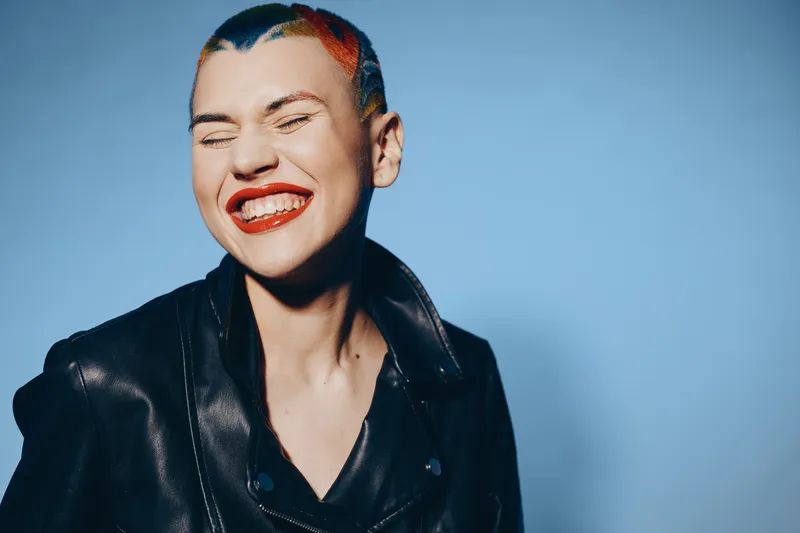Smiling woman with colorful short hair and red lipstick wearing a black leather jacket against blue background, expressing joy and bold style in studio lighting.