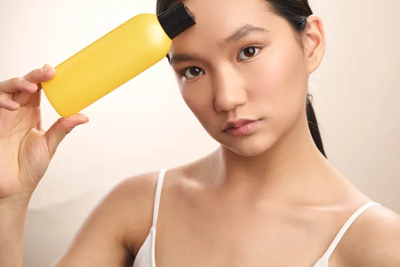asian woman holds a yellow bottle against her temple in a simple studio, presenting a calm, focused mood suitable for beauty, wellness, skincare or cosmetic product showcases.