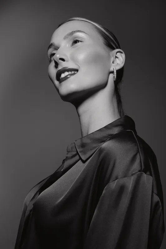 Elegant white woman with fair skin and slicked back hair poses confidently in shiny satin blouse against dark background in black and white portrait photography.