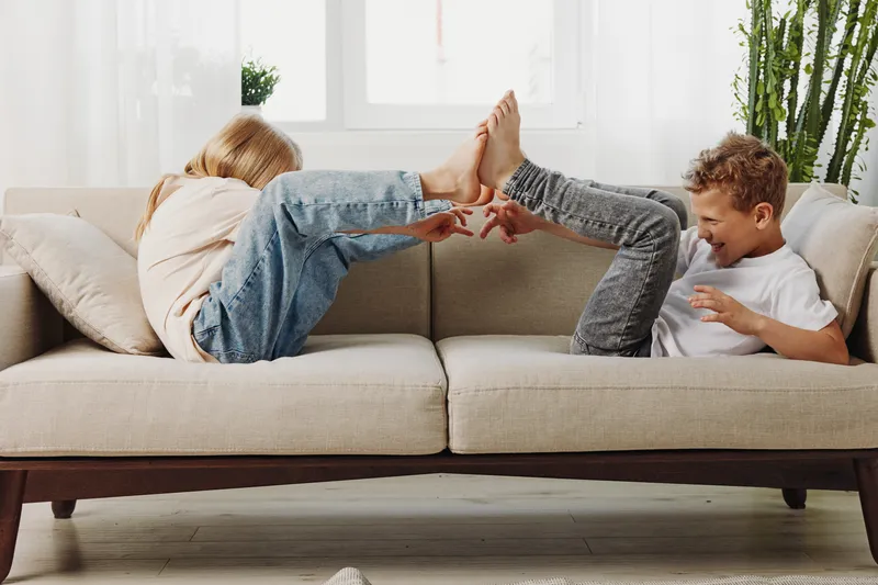 Two children playfully tickle each other lying on a beige sofa in a bright living room with natural light and green plants in the background.