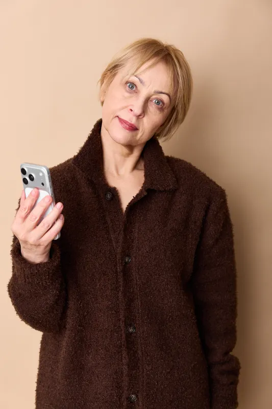 Woman smartphone portrait mature indoors beige communication technology holding studio, woman holds smartphone indoors, calm camera gaze, warm light, neutral beige background