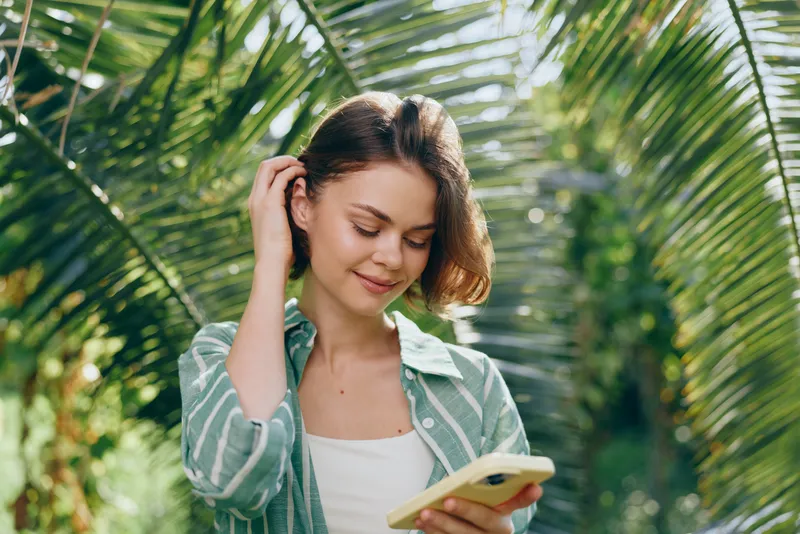 Young woman outdoors, absorbed in a tablet, surrounded by lush tropical plants; casual, serene moment of digital exploration.
