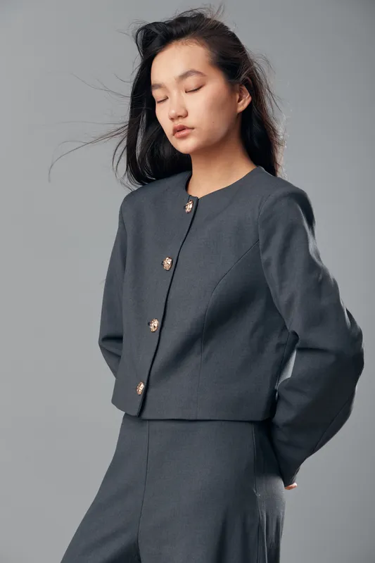 Young woman with closed eyes wearing gray formal jacket and matching trousers standing against gray studio background. Asian female model with long dark hair in minimalist fashion pose.