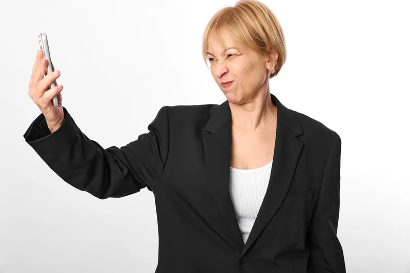 Senior woman holding smartphone in hand, studio portrait, serious expression, focused eyes on screen, white background, technology and communication concept, office business style