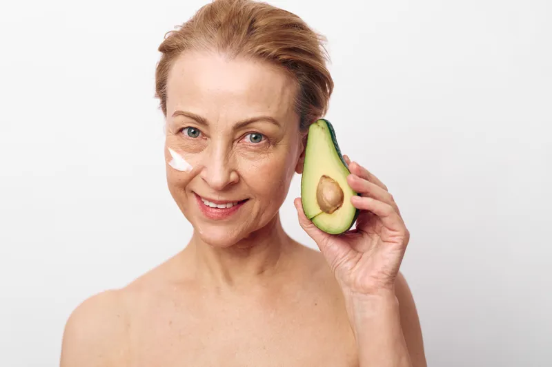 Mature woman with natural skin care cream applied on her face holds half of fresh avocado near cheek. Concept of healthy beauty, organic skincare, and natural anti aging treatment at home.