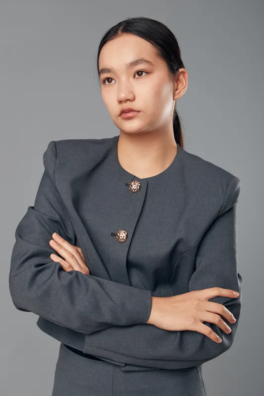 Young woman standing with crossed arms looking away in business outfit studio portrait. Female professional with serious expression in gray jacket on plain background for corporate concept.