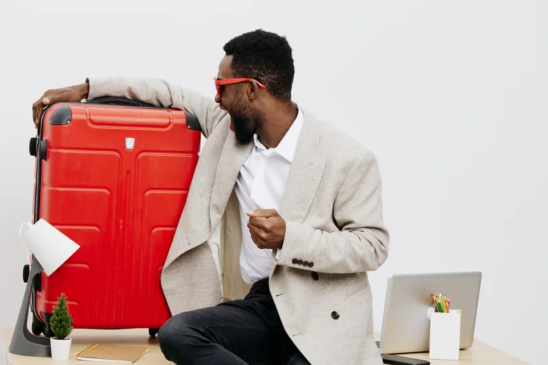 Man with suit and red glasses holding large red suitcase in modern office environment. Professional travel, business trip, airport luggage, modern lifestyle concept, casual style, workspace setup