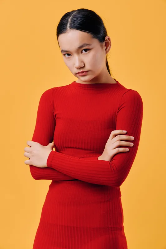 Young woman with serious face standing with crossed arms wearing red dress against yellow background in studio setting. Fashion, portrait, confident and contemplative mood for lifestyle and style