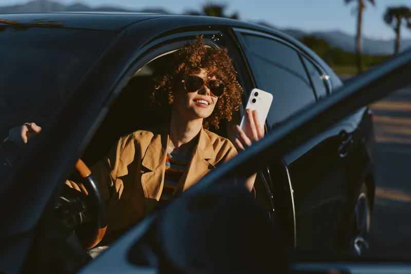 A woman sits in a car with the window down, smiling at her phone. Warm retro film colors create a casual lifestyle moment that suggests travel, freedom, and spontaneous joy.