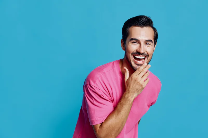 Full body smiling happy cheerful young man wear pink t-shirt looking at camera with hand touching chin isolated on plain solid blue background studio portrait. People lifestyle concept