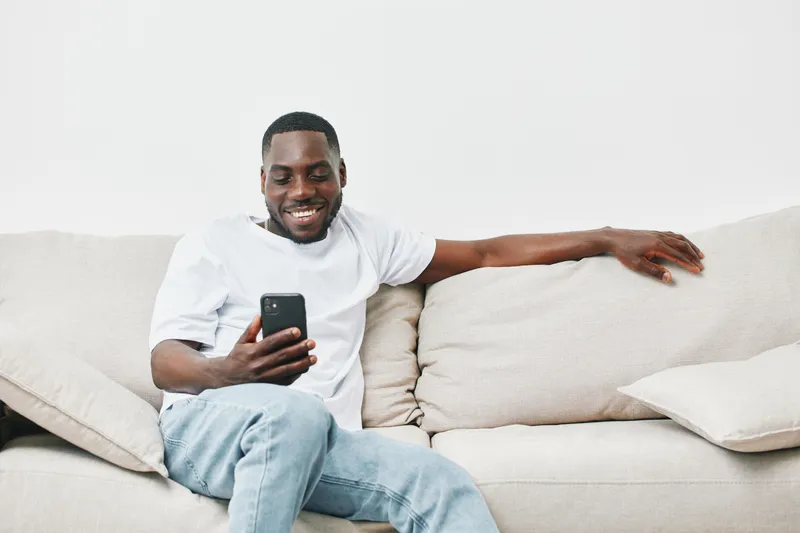 Smiling young Black man relaxing on a couch while using a smartphone, dressed casually in a white t shirt and light blue jeans, vibrant and friendly atmosphere