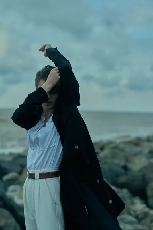 Woman on a windy beach adjusting her hair under a long coat, moody seascape with dramatic sky, a candid moment of resilience and style.