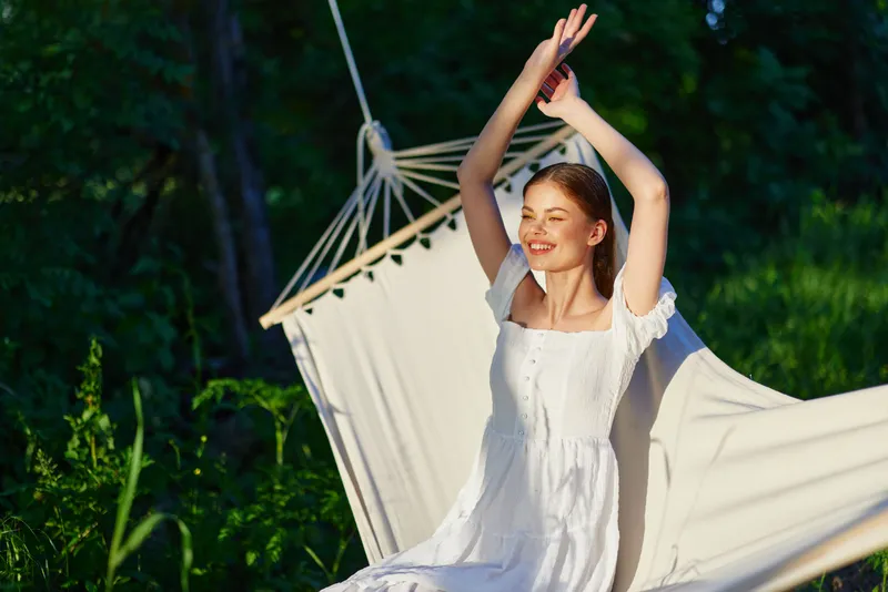 Young woman in a white dress enjoying nature, expressing joy and relaxation in a lush green setting with soft sunlight, perfect for seasonal wellness themes