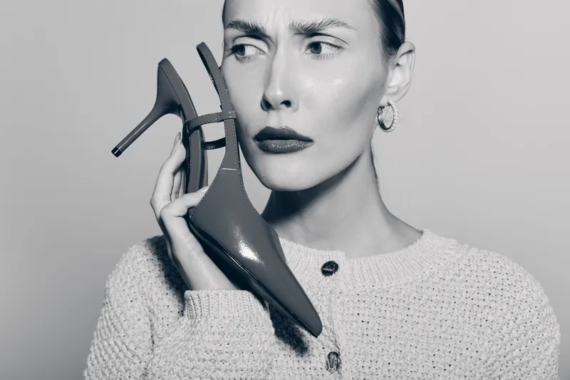 Concerned Caucasian woman with light skin holds high heel shoe near face, wearing knit sweater. Concept for fashion, emotions, and personal style.