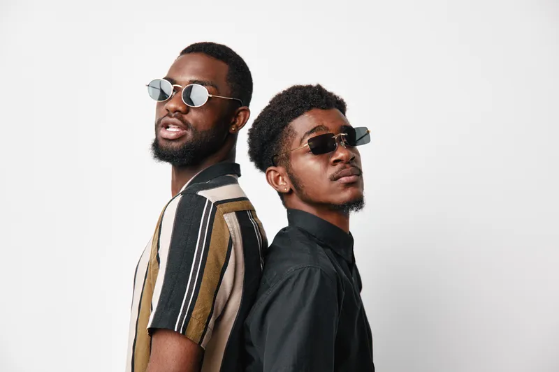 Two young African American men with dark skin wearing sunglasses and casual shirts standing back to back against white background for fashion and style concept.