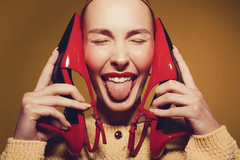 Young woman with closed eyes holds red high heels near face and sticks out tongue. Playful mood with bright shoes and cozy sweater on warm background.