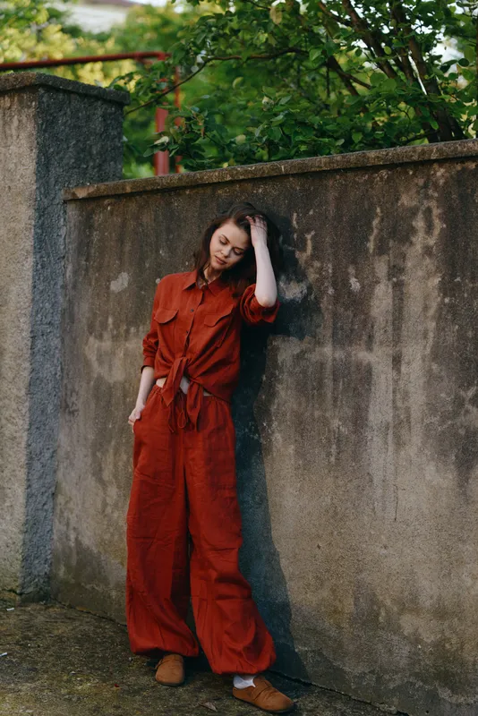 Cheerful woman outdoors in a rust red jumpsuit, leaning against a weathered wall, relaxed pose, sunlit greenery in the background, casual lifestyle moment, natural colors