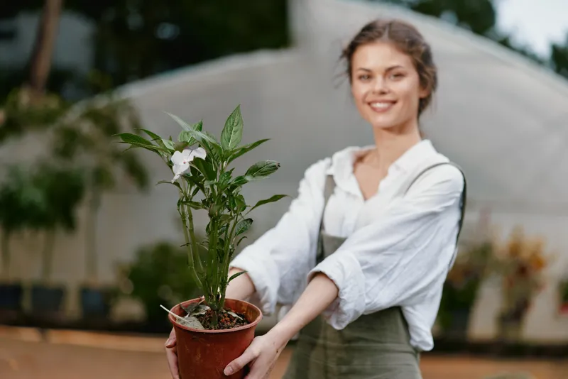 Young woman smiling while holding a potted plant in a greenhouse, portraying concepts of sustainability and gardening