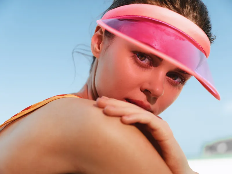 Close up portrait of a woman wearing a pink visor on a sunny day, resting her chin on her arm, with sunlit skin and a calm, playful gaze toward the camera.