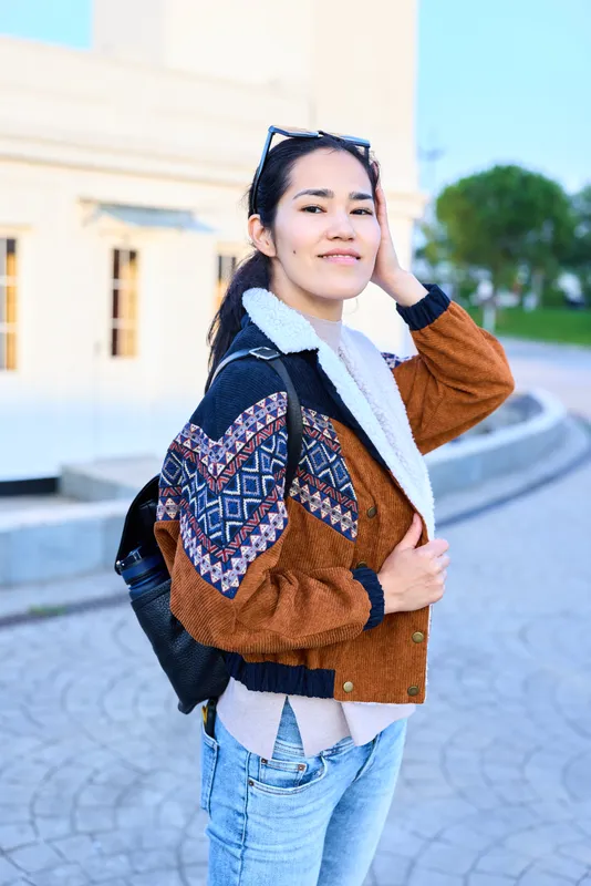 Young woman wearing a patterned jacket with denim jeans stands outdoors, backpack on, sunglasses atop head, smiling confidently in a sunny urban plaza, casual stylish vibe