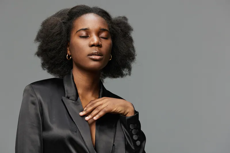 A confident African woman in a black satin blazer, showcasing self assurance with closed eyes against a neutral gray background, embodying modern elegance and empowerment