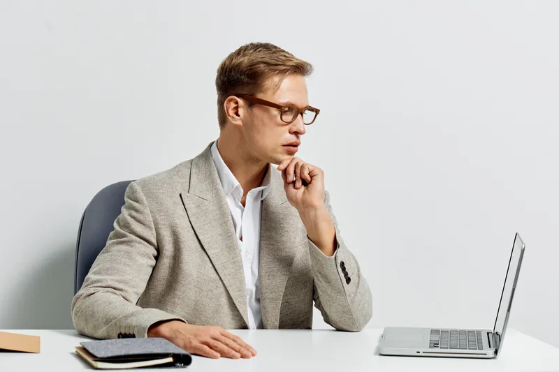 Thoughtful young man in smart casual outfit with glasses working on a laptop at desk isolated on white background. Business, technology, office, concentration, modern style, professional concept