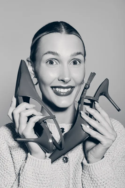 Happy Caucasian woman with fair skin holds high heel shoes near face smiling, wearing knitted sweater, studio portrait on grey background.