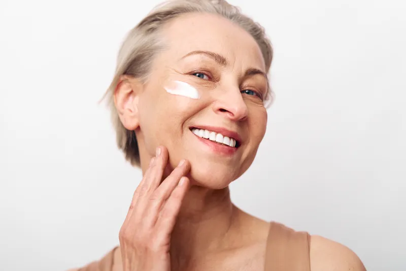 Smiling mature woman applies skincare cream on face with fingers, showing natural beauty and healthy skin on plain white background. Concept for skincare, aging, self-care, and wellness.