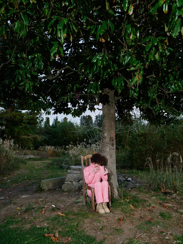 Outdoor scene with a person sitting on a chair under a leafy tree in a garden, wearing pink clothing, a serene atmosphere, natural light, a quiet moment of rest and reflection among plants.