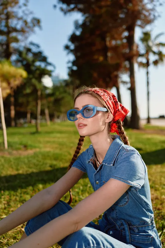 young woman wearing blue sunglasses and red bandana sitting outdoors on green grass during sunny day with trees in background
