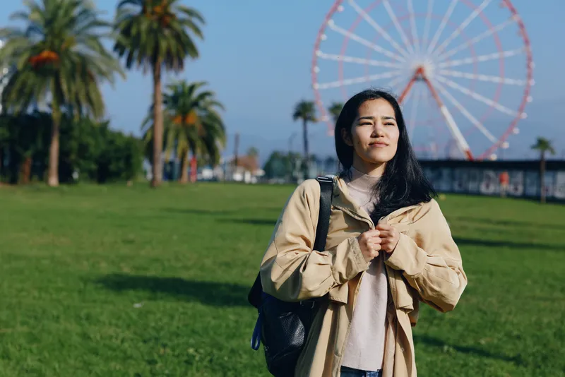 Outdoor scene with a woman in a beige jacket standing in a sunny park, palm trees and a ferris wheel in the background, conveying casual travel and relaxed moments