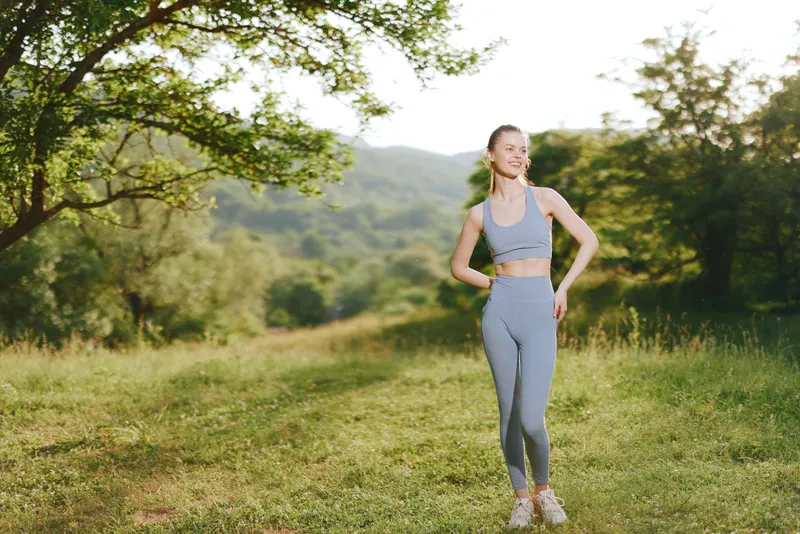 Fit young woman in gray sports attire enjoying a sunny day outdoors, representing health and fitness with a joyful expression in a natural landscape
