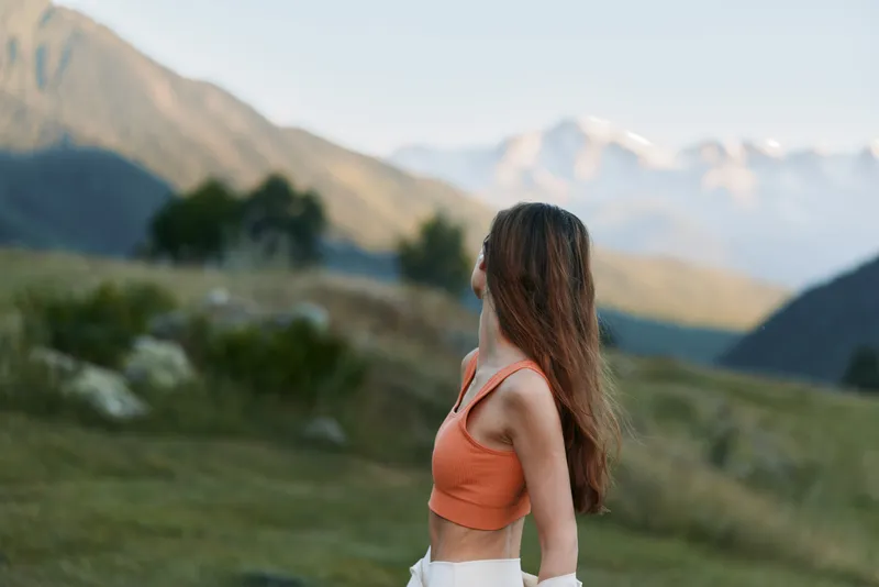 A young woman stands in a scenic mountain landscape wearing an orange top, facing away, enjoying nature, sunlight, and a peaceful outdoor moment.