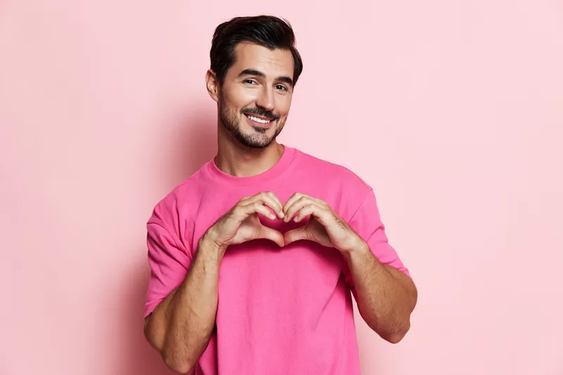 Smiling man making heart shape with hands on pink background, wearing casual bright shirt, expressing love and positivity in a vibrant studio portrait.