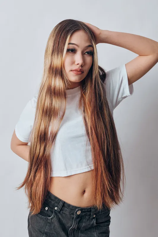 Young woman with long straight hair posing in casual white cropped t shirt and black jeans on light background. Stylish girl with natural makeup and confident expression indoors.