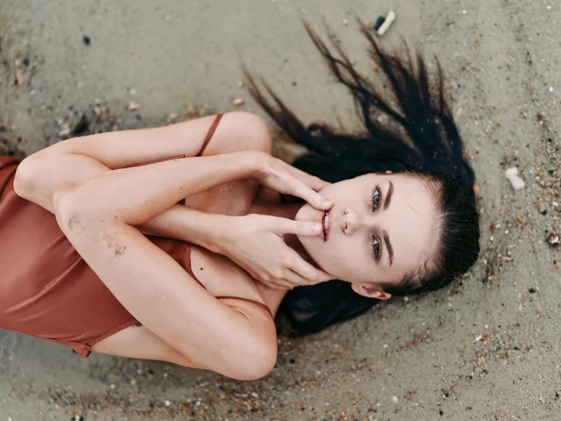 Portrait of a woman lying on sandy beach with dark hair, pale skin, and a soft gaze. The scene conveys vulnerability, calm, and tactile light across the beach surface.