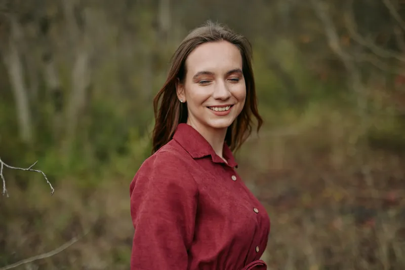 Young woman in a natural outdoor portrait wearing a red blouse, smiling with closed eyes and soft expression, capturing a genuine, authentic moment of calm confidence and honest human emotion.