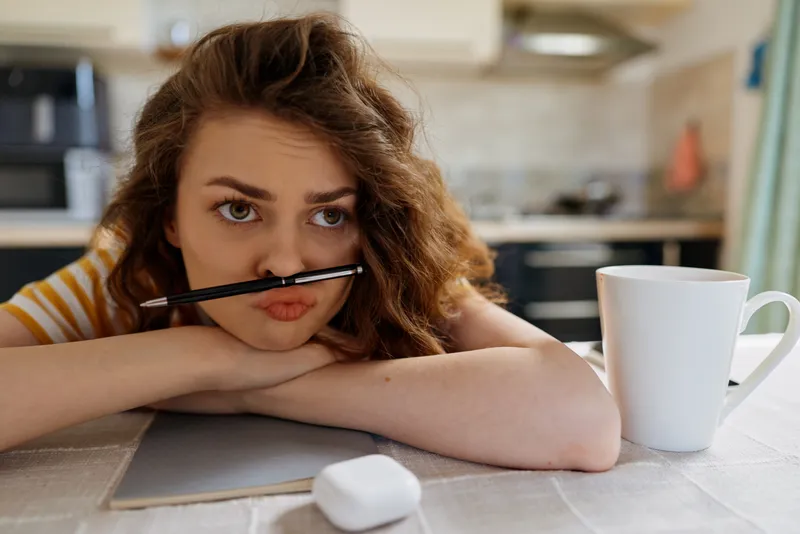 Smiling woman with curly hair resting her chin on a desk, a pencil in her mouth and a notebook nearby. Cozy home setting conveys study, creativity, and relaxed daily routines.