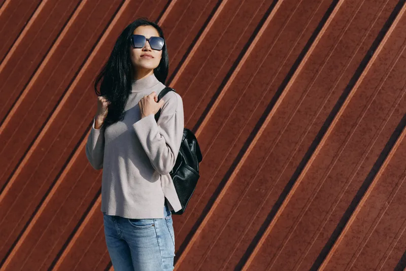 Woman with backpack, oversized sunglasses, gray sweater and jeans poses in front of a warm wooden wall, showcasing casual urban fashion and a relaxed confident attitude.