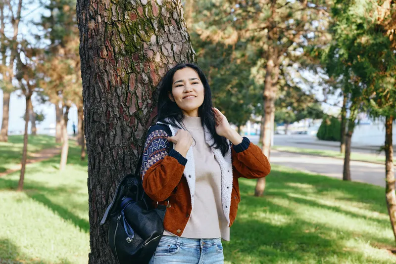 Woman leaning against a tree in a park, autumn sunlight and nature setting, casual sweater and backpack for outdoor lifestyle and travel imagery