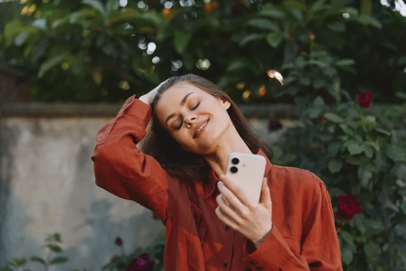 Vibrant outdoor portrait of a smiling woman in a rust top taking a selfie in a garden, showcasing relaxed lifestyle, natural light and candid joy