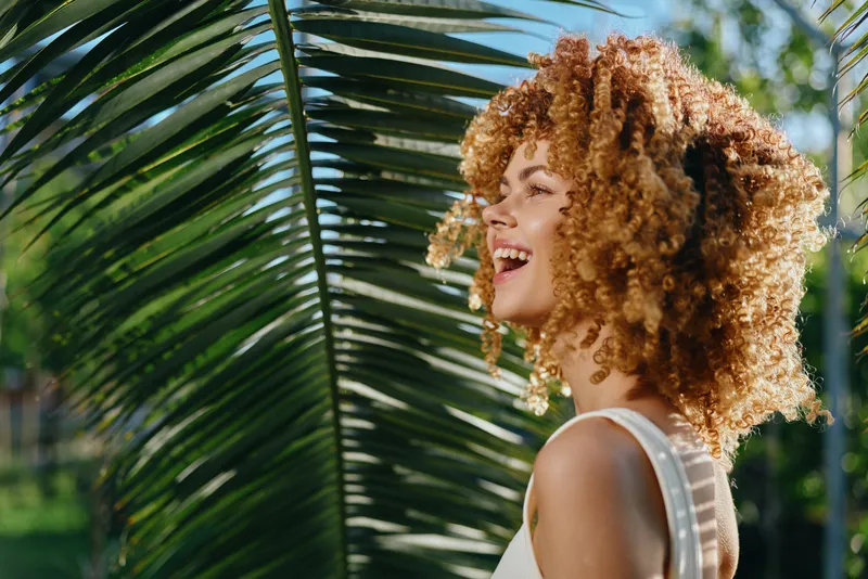 smiling woman with curly hair enjoying sunny tropical outdoors with vibrant greenery and palm leaves creating warm and joyful atmosphere