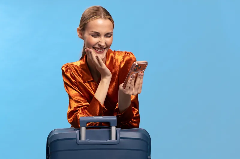 Smiling woman in orange blouse looks at smartphone while leaning on blue suitcase against light blue background. Concept of travel, communication, happy mood, and lifestyle with luggage.