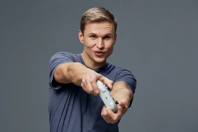 Young man holding a game controller and smiling at the camera, wearing casual gray t-shirt, isolated on plain gray background, studio portrait. People lifestyle concept